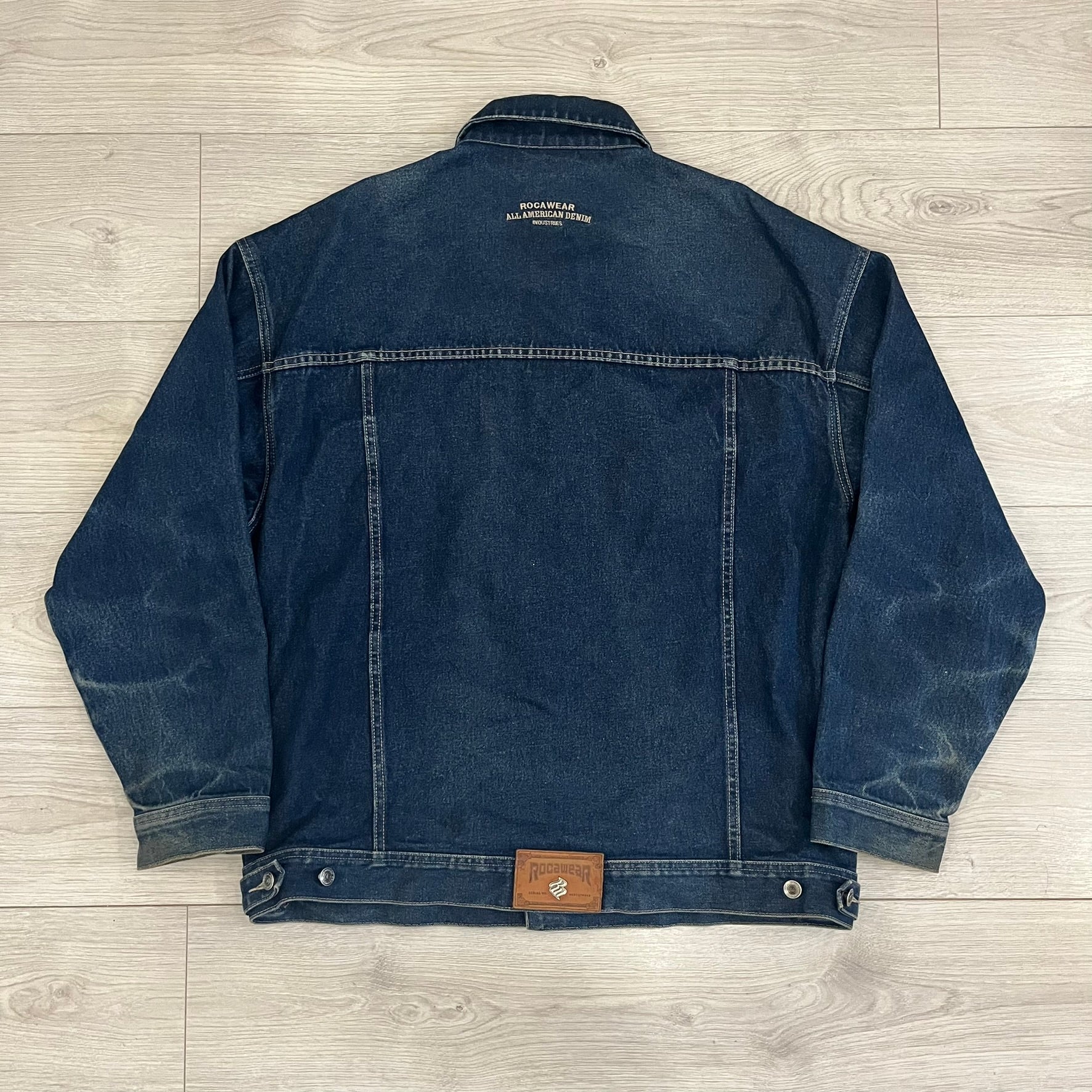 Rocawear blue denim jacket from the early 2000s, made from 100% cotton. Features an embroidered Rocawear logo on the right chest above the pocket, two buttoned chest pockets and standard side pockets. Finished with branded Rocawear metal buttons, a leather tab with metal R logo at the lower back, and “Rocawear All American Denim Industries” embroidery placed under the collar on the back. Classic structured denim fit with durable construction.