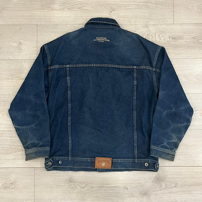 Rocawear blue denim jacket from the early 2000s, made from 100% cotton. Features an embroidered Rocawear logo on the right chest above the pocket, two buttoned chest pockets and standard side pockets. Finished with branded Rocawear metal buttons, a leather tab with metal R logo at the lower back, and “Rocawear All American Denim Industries” embroidery placed under the collar on the back. Classic structured denim fit with durable construction.