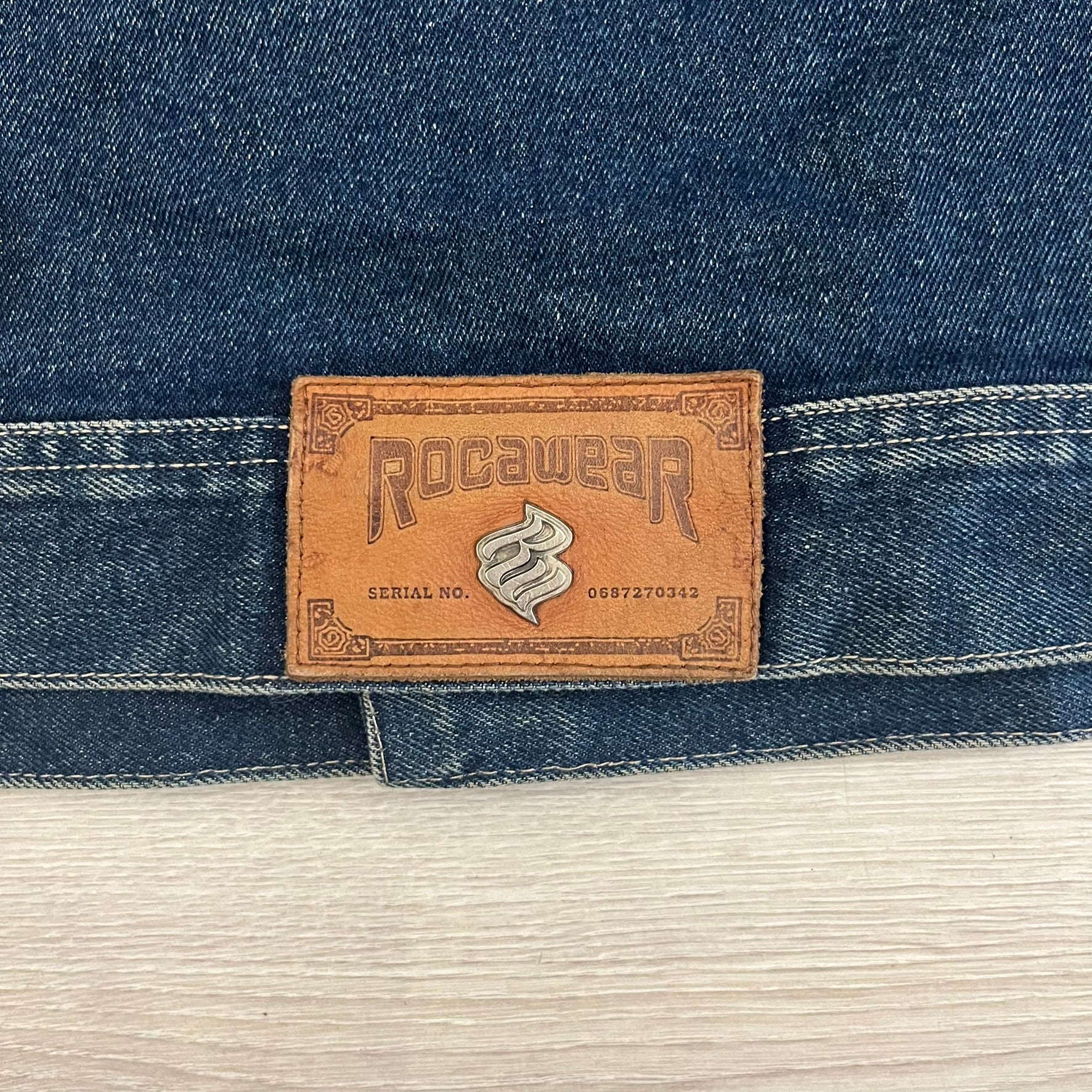 Rocawear blue denim jacket from the early 2000s, made from 100% cotton. Features an embroidered Rocawear logo on the right chest above the pocket, two buttoned chest pockets and standard side pockets. Finished with branded Rocawear metal buttons, a leather tab with metal R logo at the lower back, and “Rocawear All American Denim Industries” embroidery placed under the collar on the back. Classic structured denim fit with durable construction.