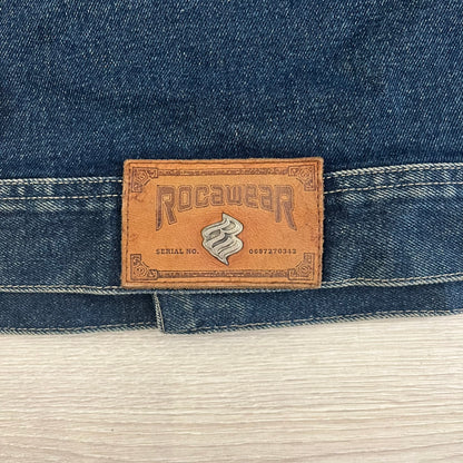 Rocawear blue denim jacket from the early 2000s, made from 100% cotton. Features an embroidered Rocawear logo on the right chest above the pocket, two buttoned chest pockets and standard side pockets. Finished with branded Rocawear metal buttons, a leather tab with metal R logo at the lower back, and “Rocawear All American Denim Industries” embroidery placed under the collar on the back. Classic structured denim fit with durable construction.