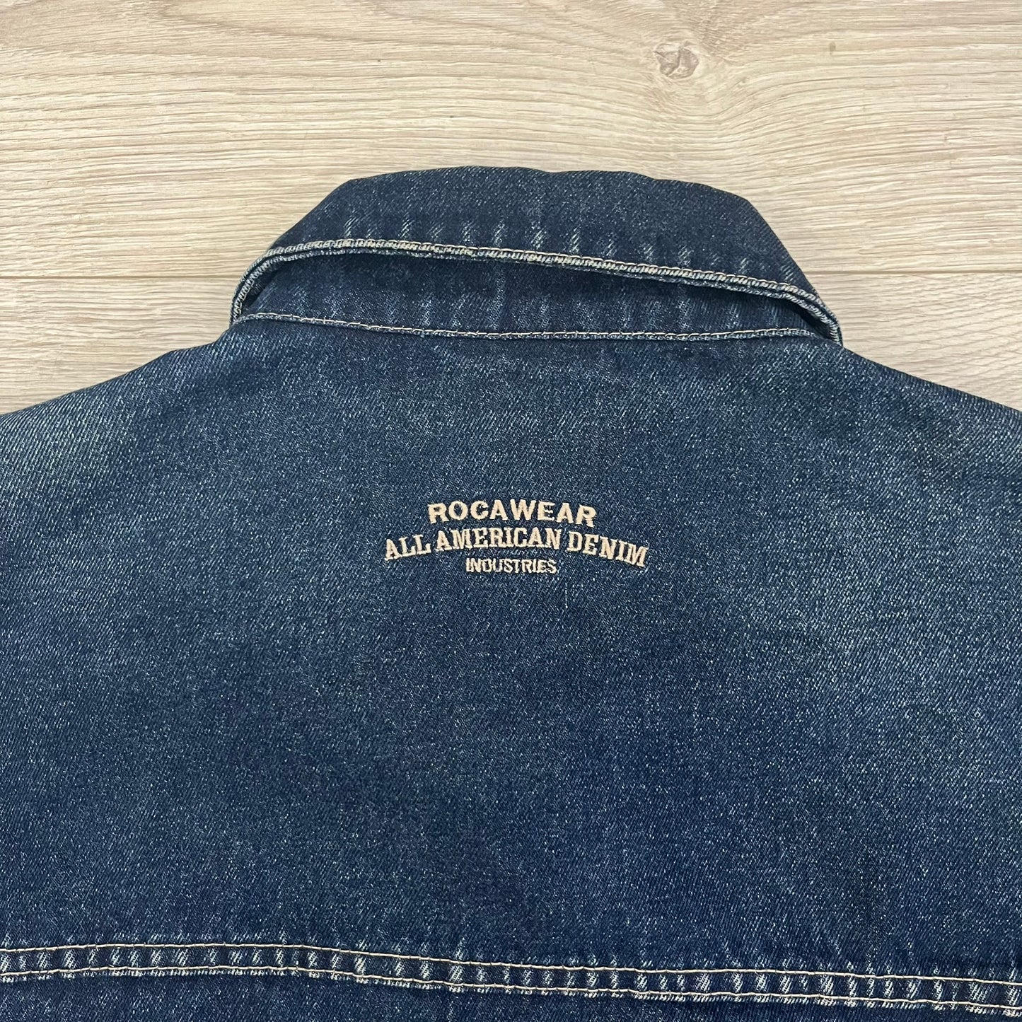 Rocawear blue denim jacket from the early 2000s, made from 100% cotton. Features an embroidered Rocawear logo on the right chest above the pocket, two buttoned chest pockets and standard side pockets. Finished with branded Rocawear metal buttons, a leather tab with metal R logo at the lower back, and “Rocawear All American Denim Industries” embroidery placed under the collar on the back. Classic structured denim fit with durable construction.