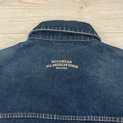 Rocawear blue denim jacket from the early 2000s, made from 100% cotton. Features an embroidered Rocawear logo on the right chest above the pocket, two buttoned chest pockets and standard side pockets. Finished with branded Rocawear metal buttons, a leather tab with metal R logo at the lower back, and “Rocawear All American Denim Industries” embroidery placed under the collar on the back. Classic structured denim fit with durable construction.