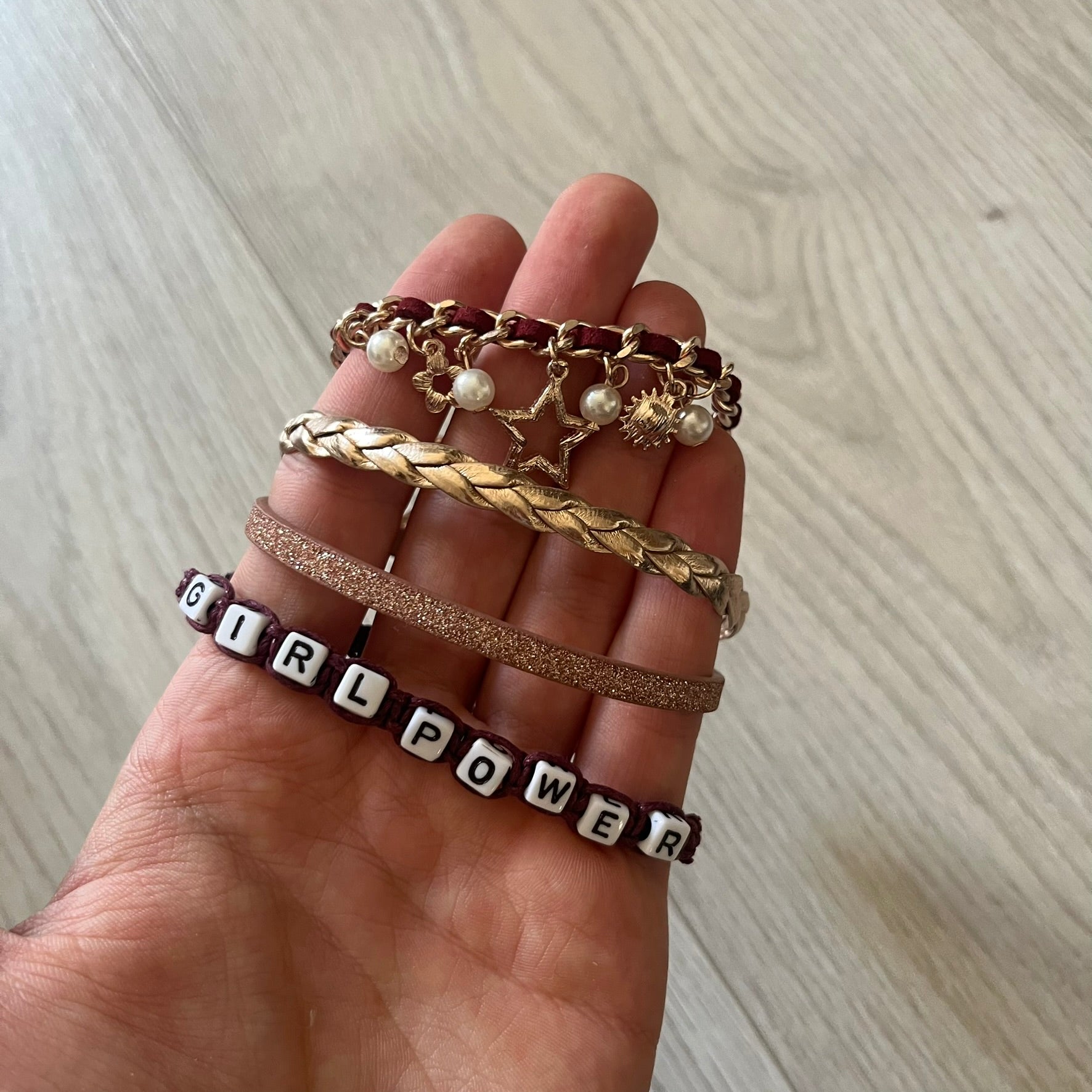 Set of four Rocawear bracelets, each with distinct design elements. Highlights include a “Girl Power” cube bracelet, a burgundy string with gold chain and pearl/zirconia charms, and two simple rose gold bands. Comes presented in a holographic silver box, ideal for collectors or as a stylish accessory set.