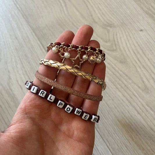 Set of four Rocawear bracelets, each with distinct design elements. Highlights include a “Girl Power” cube bracelet, a burgundy string with gold chain and pearl/zirconia charms, and two simple rose gold bands. Comes presented in a holographic silver box, ideal for collectors or as a stylish accessory set.