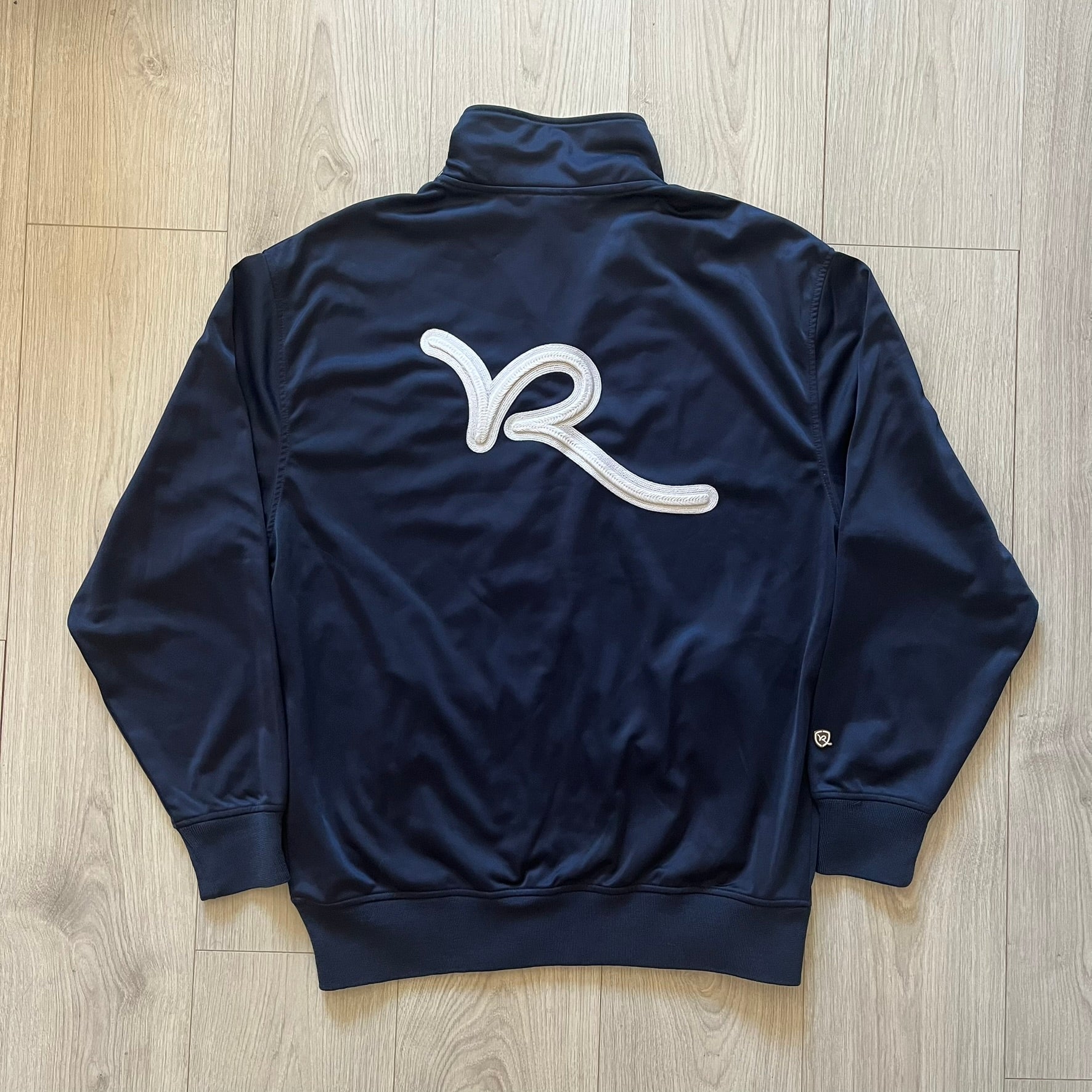 Rocawear Dark Navy Vintage Track Jacket, featuring a small embroidered logo on the chest and a large white logo across the back, finished with a silver emblem near the wrist and a detailed silver zipper.