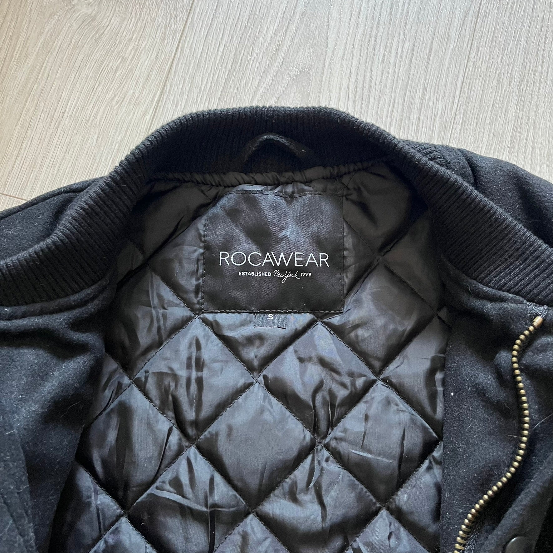 Rocawear black bomber jacket detailed with multiple statement patches, including “R” and “W” on the shoulders, a Rocawear logo with a “4” on the chest, and a large text logo across the back. Crafted with great detailing, it reflects the brand’s early 2000s streetwear identity.