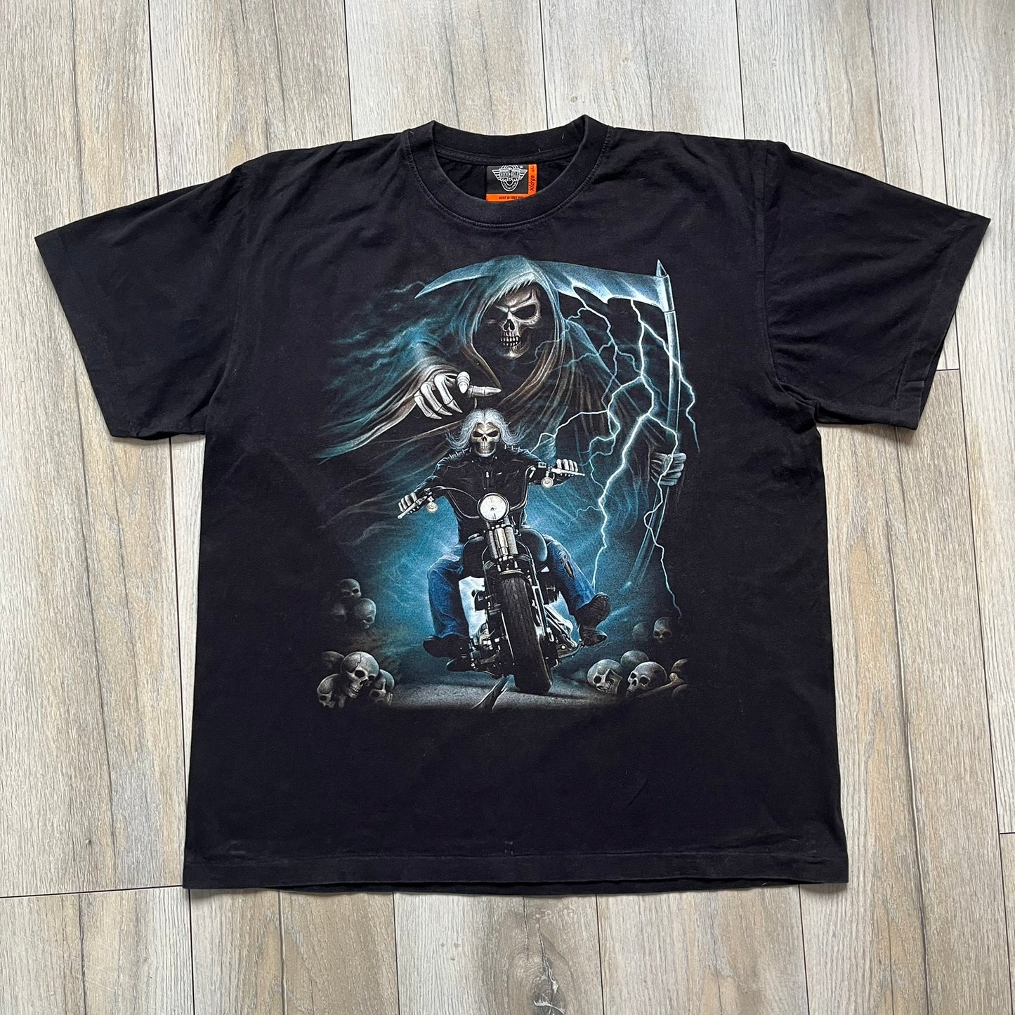 Rock Eagle 2010s Skull Biker Tee is a black T-shirt featuring a large front graphic of a skull-faced biker riding a motorcycle while being chased by a skeletal Grim Reaper holding a scythe. The artwork includes lightning strikes, skulls, and detailed airbrush-style effects throughout the scene. The back features a graphic of a winged engine with a skull at the center. Finished with a Rock Eagle neck tag.
