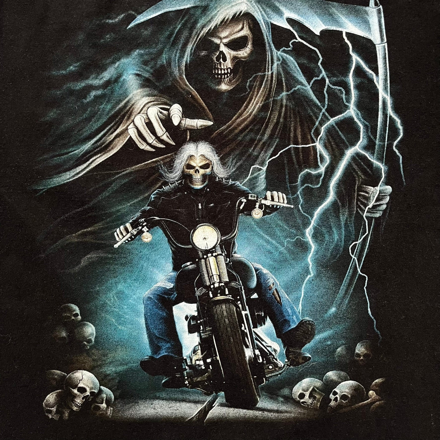 Rock Eagle 2010s Skull Biker Tee is a black T-shirt featuring a large front graphic of a skull-faced biker riding a motorcycle while being chased by a skeletal Grim Reaper holding a scythe. The artwork includes lightning strikes, skulls, and detailed airbrush-style effects throughout the scene. The back features a graphic of a winged engine with a skull at the center. Finished with a Rock Eagle neck tag.
