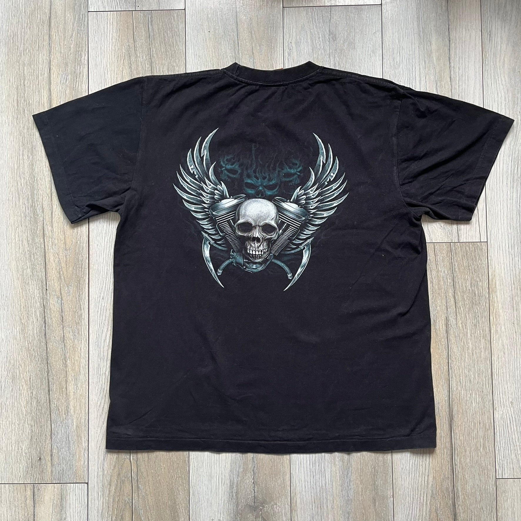 Rock Eagle 2010s Skull Biker Tee is a black T-shirt featuring a large front graphic of a skull-faced biker riding a motorcycle while being chased by a skeletal Grim Reaper holding a scythe. The artwork includes lightning strikes, skulls, and detailed airbrush-style effects throughout the scene. The back features a graphic of a winged engine with a skull at the center. Finished with a Rock Eagle neck tag.
