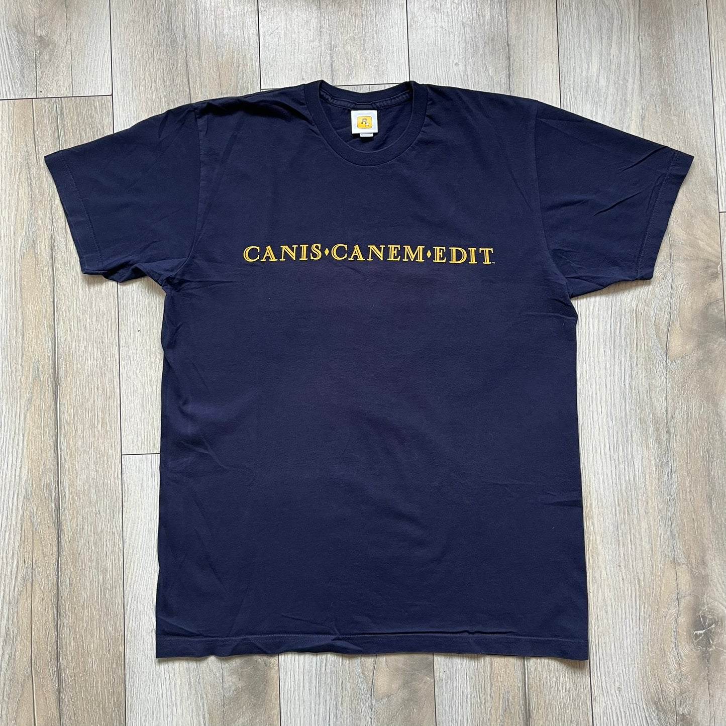 Rockstar Games 'Canis Canem Edit (Bully)' Navy Tee is a dark navy T-shirt made in the USA featuring a yellow "Canis Canem Edit" text on the chest, referencing the original Bully game title. The back displays a large yellow Bullworth Academy logo, while a small yellow Rockstar logo is printed on the sleeve. The shirt includes an official Rockstar Games neck tag.