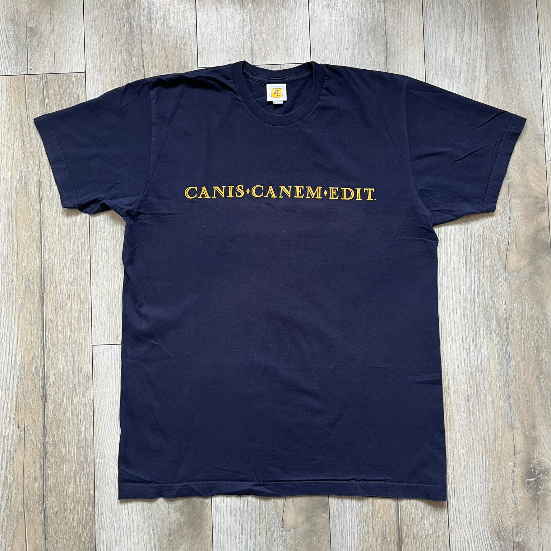 Rockstar Games 'Canis Canem Edit (Bully)' Navy Tee is a dark navy T-shirt made in the USA featuring a yellow "Canis Canem Edit" text on the chest, referencing the original Bully game title. The back displays a large yellow Bullworth Academy logo, while a small yellow Rockstar logo is printed on the sleeve. The shirt includes an official Rockstar Games neck tag.