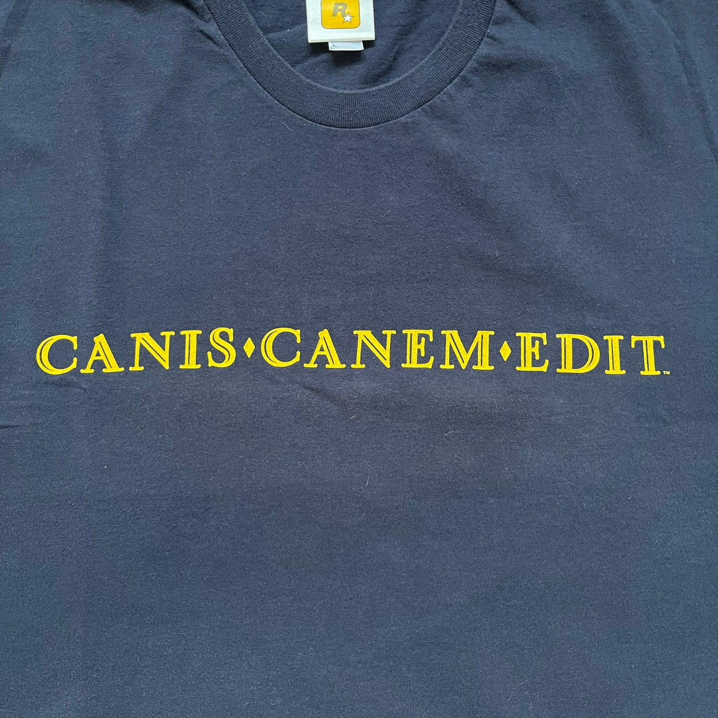 Rockstar Games 'Canis Canem Edit (Bully)' Navy Tee is a dark navy T-shirt made in the USA featuring a yellow "Canis Canem Edit" text on the chest, referencing the original Bully game title. The back displays a large yellow Bullworth Academy logo, while a small yellow Rockstar logo is printed on the sleeve. The shirt includes an official Rockstar Games neck tag.