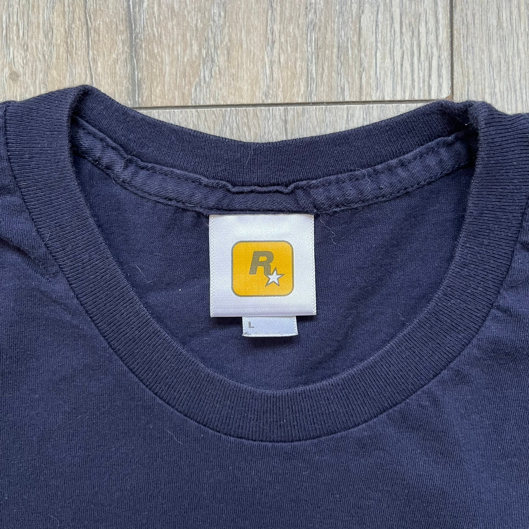 Rockstar Games 'Canis Canem Edit (Bully)' Navy Tee is a dark navy T-shirt made in the USA featuring a yellow "Canis Canem Edit" text on the chest, referencing the original Bully game title. The back displays a large yellow Bullworth Academy logo, while a small yellow Rockstar logo is printed on the sleeve. The shirt includes an official Rockstar Games neck tag.