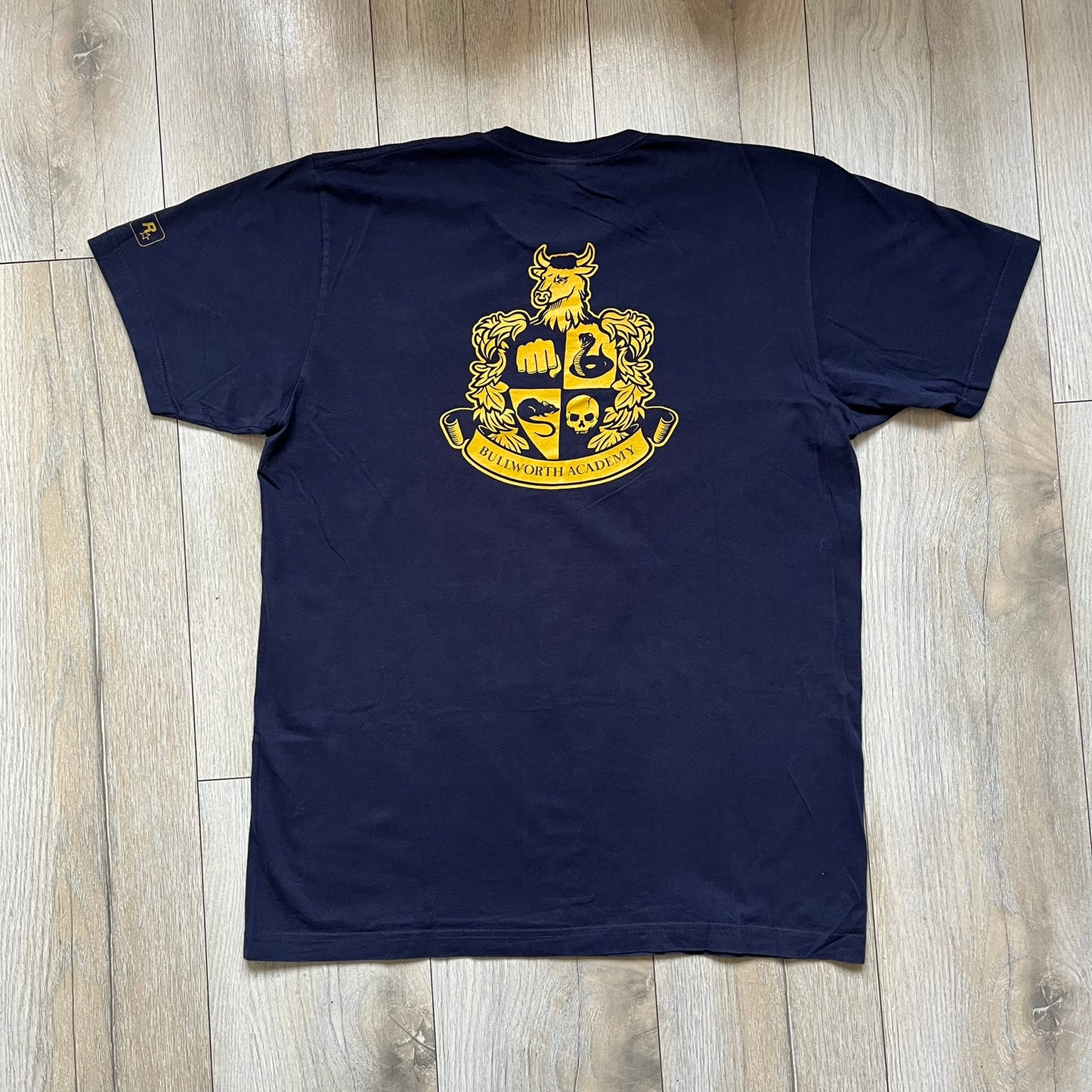 Rockstar Games 'Canis Canem Edit (Bully)' Navy Tee is a dark navy T-shirt made in the USA featuring a yellow "Canis Canem Edit" text on the chest, referencing the original Bully game title. The back displays a large yellow Bullworth Academy logo, while a small yellow Rockstar logo is printed on the sleeve. The shirt includes an official Rockstar Games neck tag.