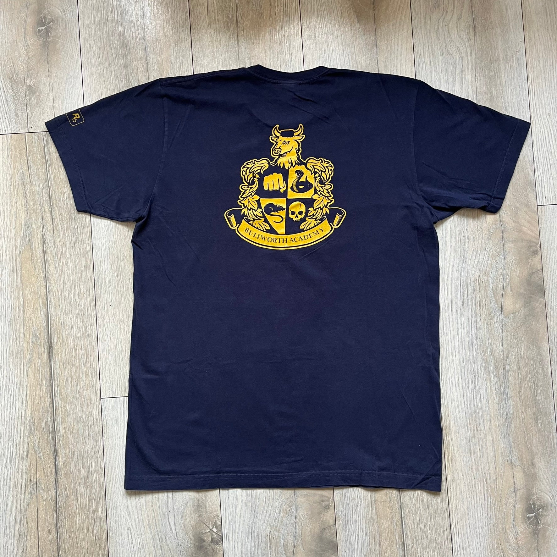 Rockstar Games 'Canis Canem Edit (Bully)' Navy Tee is a dark navy T-shirt made in the USA featuring a yellow "Canis Canem Edit" text on the chest, referencing the original Bully game title. The back displays a large yellow Bullworth Academy logo, while a small yellow Rockstar logo is printed on the sleeve. The shirt includes an official Rockstar Games neck tag.