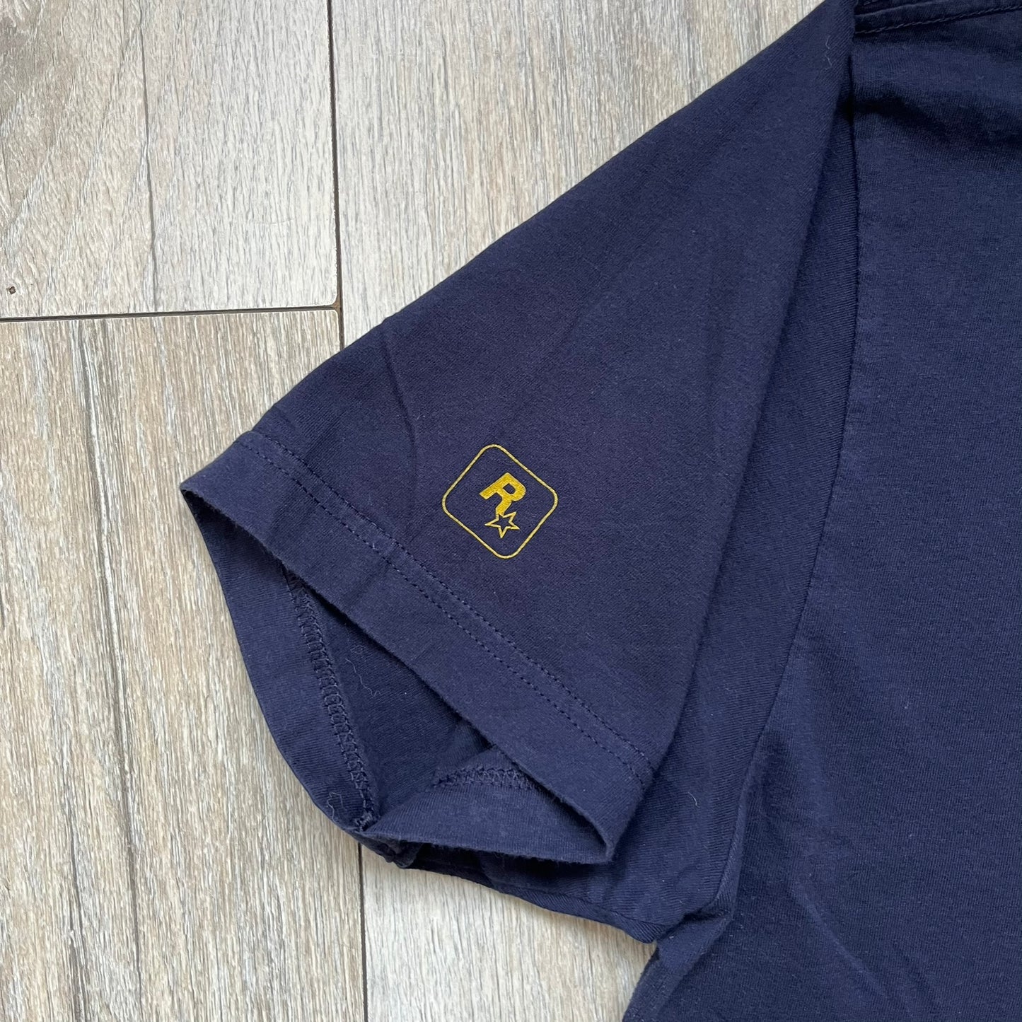 Rockstar Games 'Canis Canem Edit (Bully)' Navy Tee is a dark navy T-shirt made in the USA featuring a yellow "Canis Canem Edit" text on the chest, referencing the original Bully game title. The back displays a large yellow Bullworth Academy logo, while a small yellow Rockstar logo is printed on the sleeve. The shirt includes an official Rockstar Games neck tag.