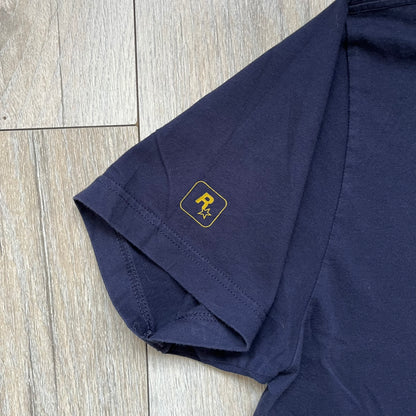 Rockstar Games 'Canis Canem Edit (Bully)' Navy Tee is a dark navy T-shirt made in the USA featuring a yellow "Canis Canem Edit" text on the chest, referencing the original Bully game title. The back displays a large yellow Bullworth Academy logo, while a small yellow Rockstar logo is printed on the sleeve. The shirt includes an official Rockstar Games neck tag.