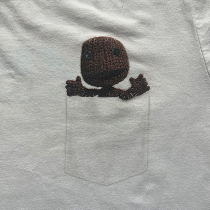 Sackboy: A Big Adventure 2020 White Promo Tee is a white cotton T-shirt featuring official Sony licensing and a “not for sale” internal tag. The front includes a small printed faux chest pocket design with Sackboy waving from inside it. The upper back displays a large “Sackboy: A Big Adventure” game logo print.