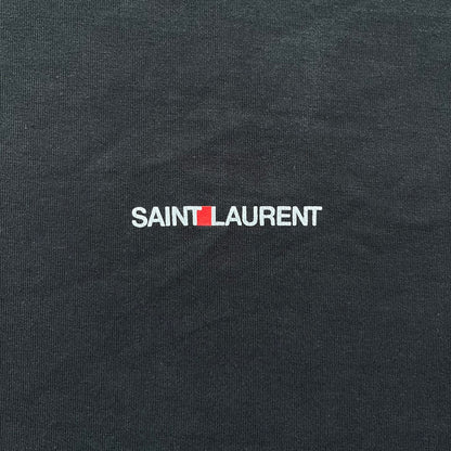 Saint Laurent 2018 Black Logo Tee is a black short-sleeve T-shirt made in Italy from 100% cotton. The chest features a small logo print with white “Saint,” a red square, and white “Laurent” detailing. The shirt has a slightly elongated front hem compared to the back.