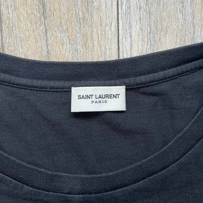 Saint Laurent 2018 Black Logo Tee is a black short-sleeve T-shirt made in Italy from 100% cotton. The chest features a small logo print with white “Saint,” a red square, and white “Laurent” detailing. The shirt has a slightly elongated front hem compared to the back.