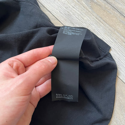 Saint Laurent 2018 Black Logo Tee is a black short-sleeve T-shirt made in Italy from 100% cotton. The chest features a small logo print with white “Saint,” a red square, and white “Laurent” detailing. The shirt has a slightly elongated front hem compared to the back.