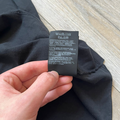 Saint Laurent 2018 Black Logo Tee is a black short-sleeve T-shirt made in Italy from 100% cotton. The chest features a small logo print with white “Saint,” a red square, and white “Laurent” detailing. The shirt has a slightly elongated front hem compared to the back.