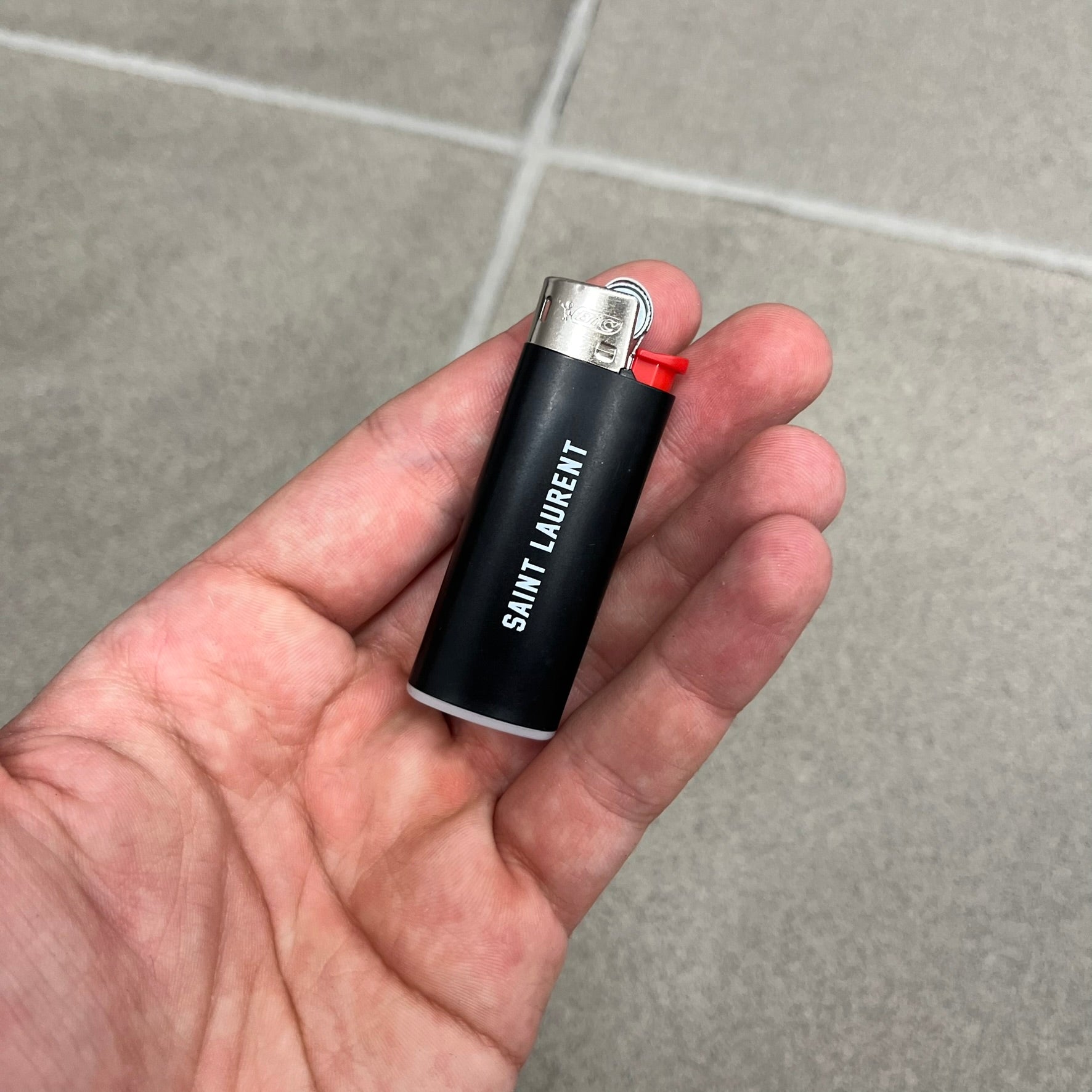 Saint Laurent Black 213 Saint-Honore Lighter is a black BIC gas lighter featuring white Saint Laurent branding on one side and 213 Saint-Honore Paris on the other side. It is an in-store-only item made in Spain.
