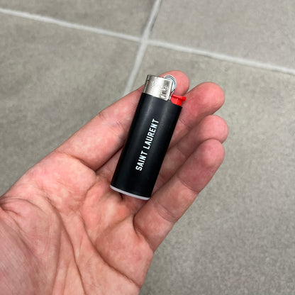 Saint Laurent Black 213 Saint-Honore Lighter is a black BIC gas lighter featuring white Saint Laurent branding on one side and 213 Saint-Honore Paris on the other side. It is an in-store-only item made in Spain.
