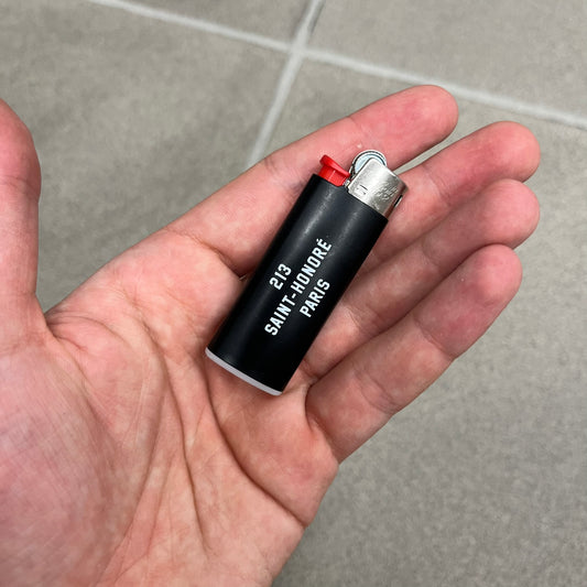 Saint Laurent Black 213 Saint-Honore Lighter is a black BIC gas lighter featuring white Saint Laurent branding on one side and 213 Saint-Honore Paris on the other side. It is an in-store-only item made in Spain.
