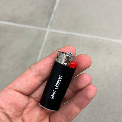 Saint Laurent Black 213 Saint-Honore Lighter is a black BIC gas lighter featuring white Saint Laurent branding on one side and 213 Saint-Honore Paris on the other side. It is an in-store-only item made in Spain.
