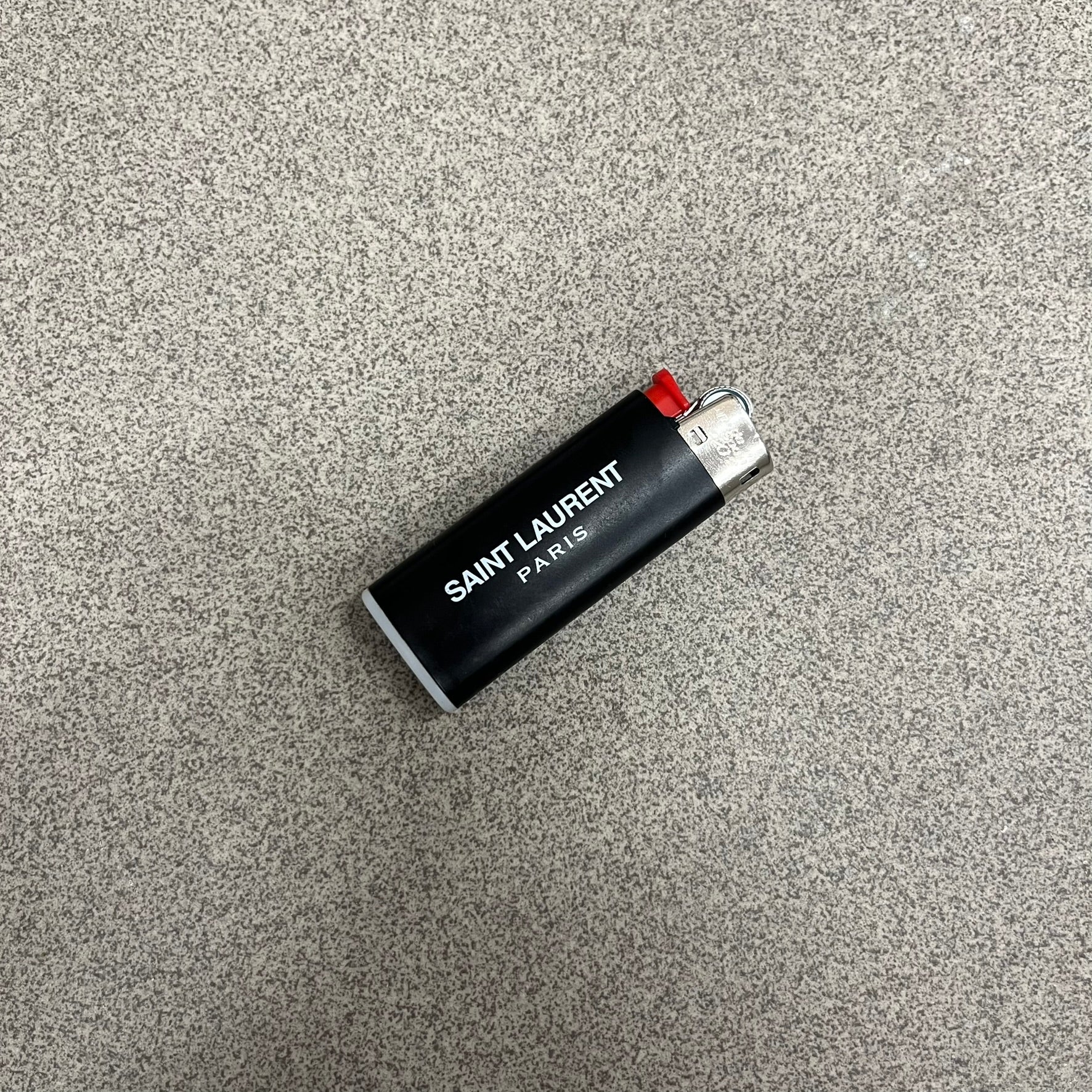 Saint Laurent Black 213 Saint-Honore Lighter is a black BIC gas lighter featuring white Saint Laurent branding on one side and Appelle Moi text on the other. It is an in-store-only item made in Spain.
