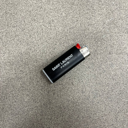Saint Laurent Black 213 Saint-Honore Lighter is a black BIC gas lighter featuring white Saint Laurent branding on one side and Appelle Moi text on the other. It is an in-store-only item made in Spain.
