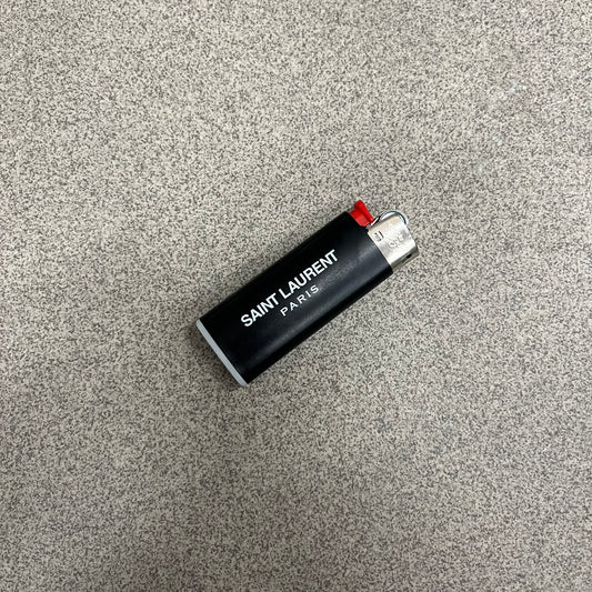 Saint Laurent Black 213 Saint-Honore Lighter is a black BIC gas lighter featuring white Saint Laurent branding on one side and Appelle Moi text on the other. It is an in-store-only item made in Spain.

