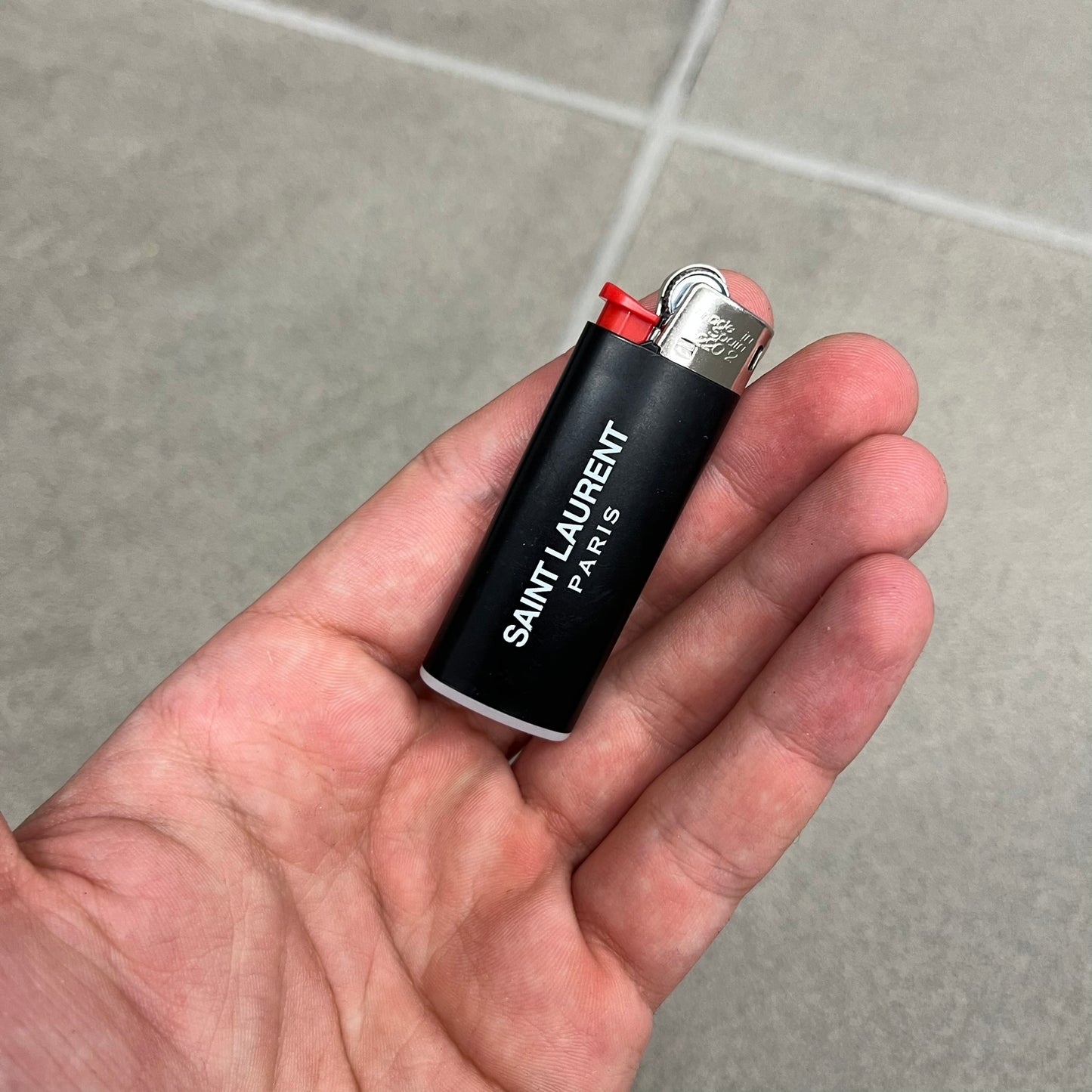 Saint Laurent Black 213 Saint-Honore Lighter is a black BIC gas lighter featuring white Saint Laurent branding on one side and Appelle Moi text on the other. It is an in-store-only item made in Spain.
