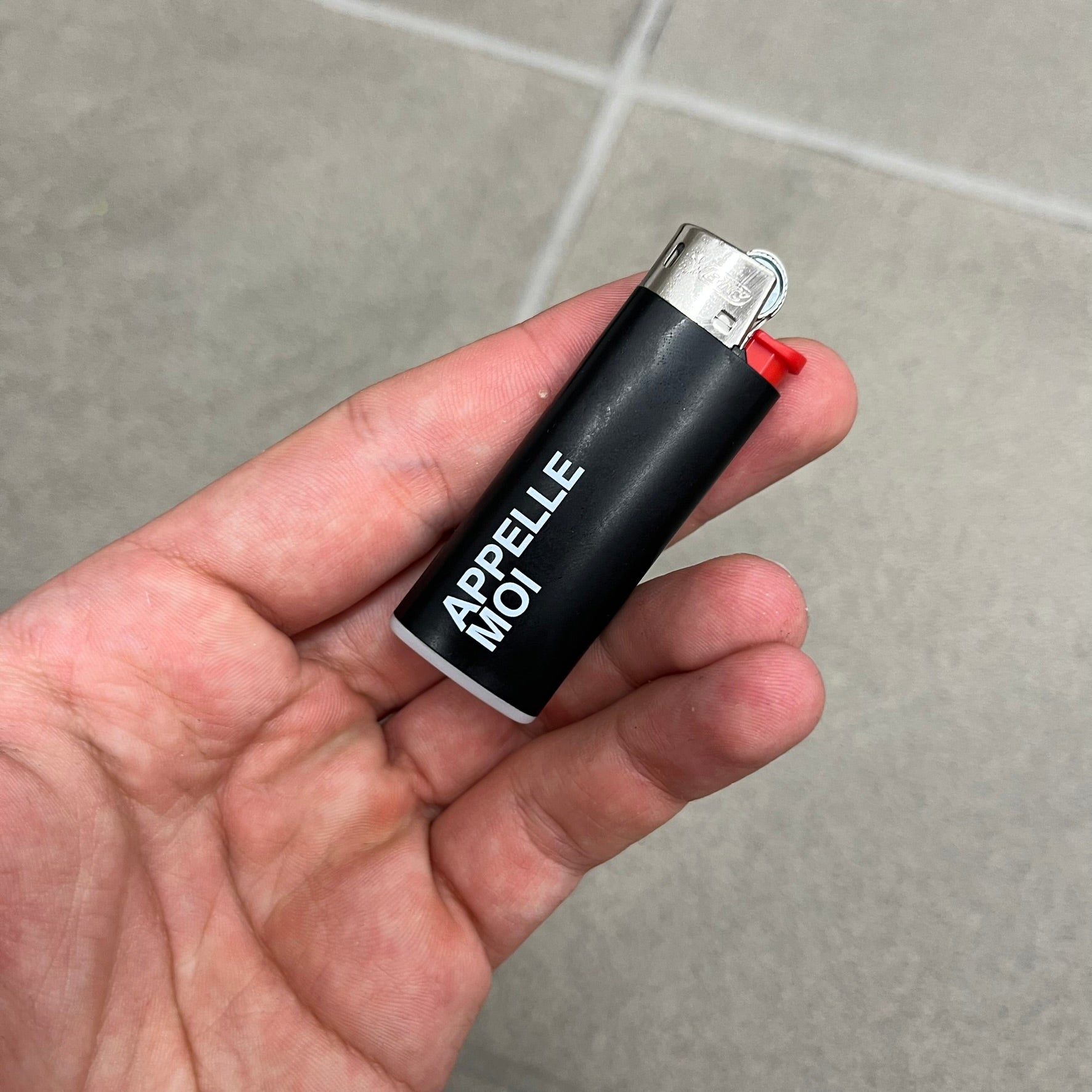 Saint Laurent Black 213 Saint-Honore Lighter is a black BIC gas lighter featuring white Saint Laurent branding on one side and Appelle Moi text on the other. It is an in-store-only item made in Spain.
