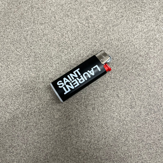  Saint Laurent Black Logo Lighter is a white BIC gas lighter featuring white Saint Laurent branding, with “Saint” printed above and “Laurent” mirrored underneath. It is an in-store-only item made in Spain. 