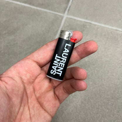  Saint Laurent Black Logo Lighter is a white BIC gas lighter featuring white Saint Laurent branding, with “Saint” printed above and “Laurent” mirrored underneath. It is an in-store-only item made in Spain. 