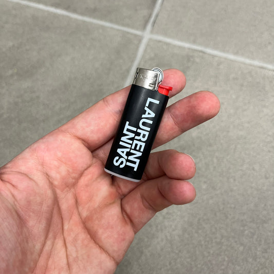  Saint Laurent Black Logo Lighter is a white BIC gas lighter featuring white Saint Laurent branding, with “Saint” printed above and “Laurent” mirrored underneath. It is an in-store-only item made in Spain. 