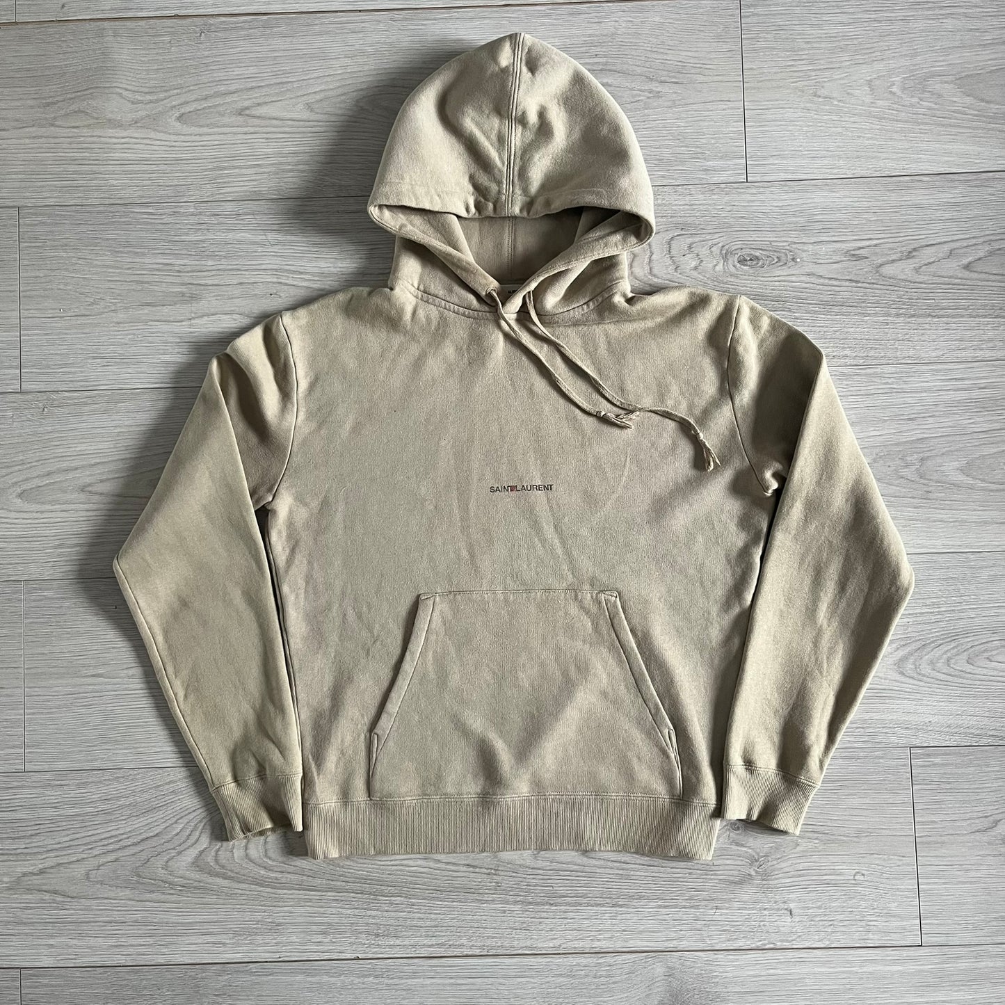 Crafted in France from 100% cotton, this Saint Laurent Beige Hoodie features a minimalist aesthetic with the brand’s signature small Saint Laurent chest logo and red square detail. Designed with a classic front pocket and clean drawstrings without metal tips, it delivers understated Parisian luxury in a neutral tone.