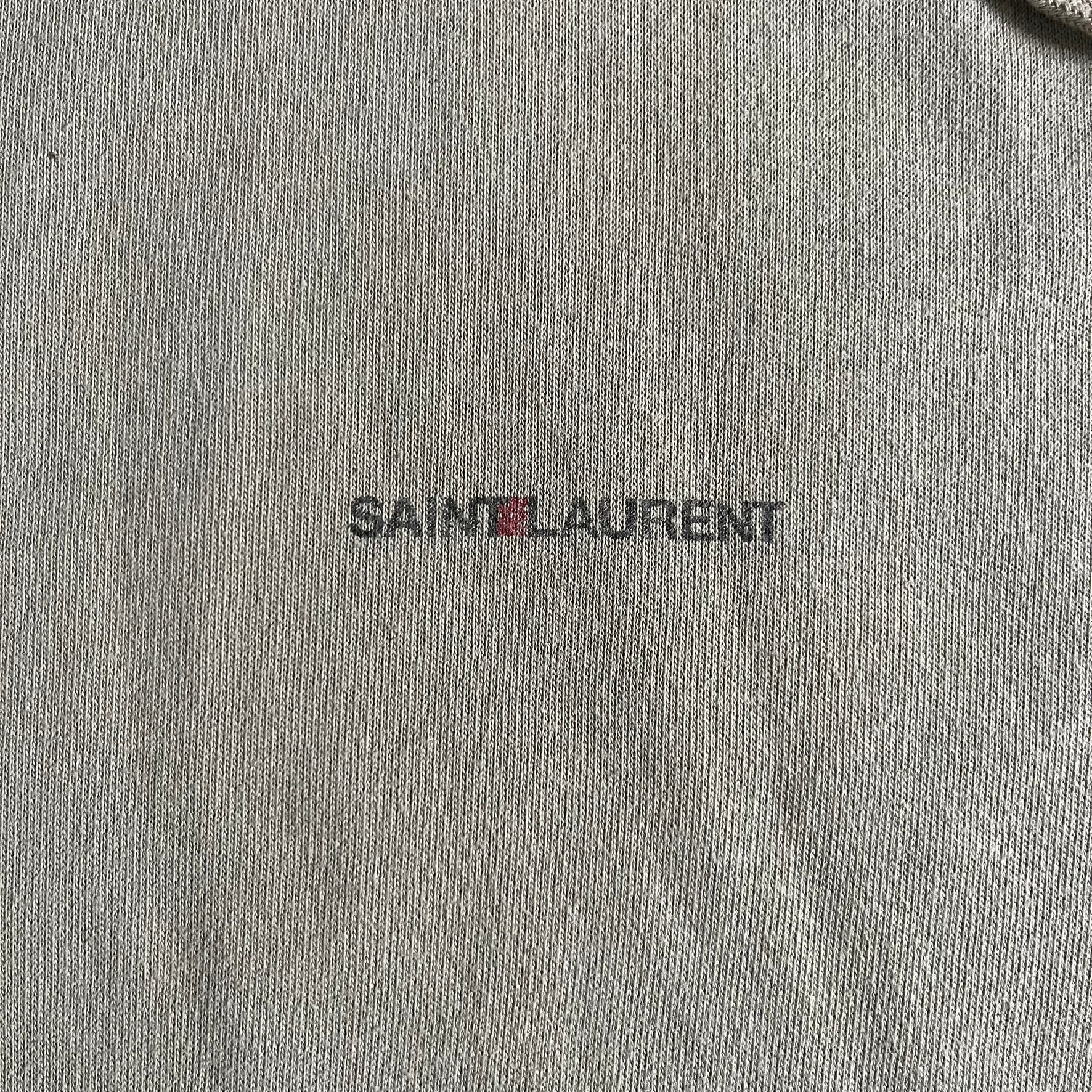 Crafted in France from 100% cotton, this Saint Laurent Beige Hoodie features a minimalist aesthetic with the brand’s signature small Saint Laurent chest logo and red square detail. Designed with a classic front pocket and clean drawstrings without metal tips, it delivers understated Parisian luxury in a neutral tone.
