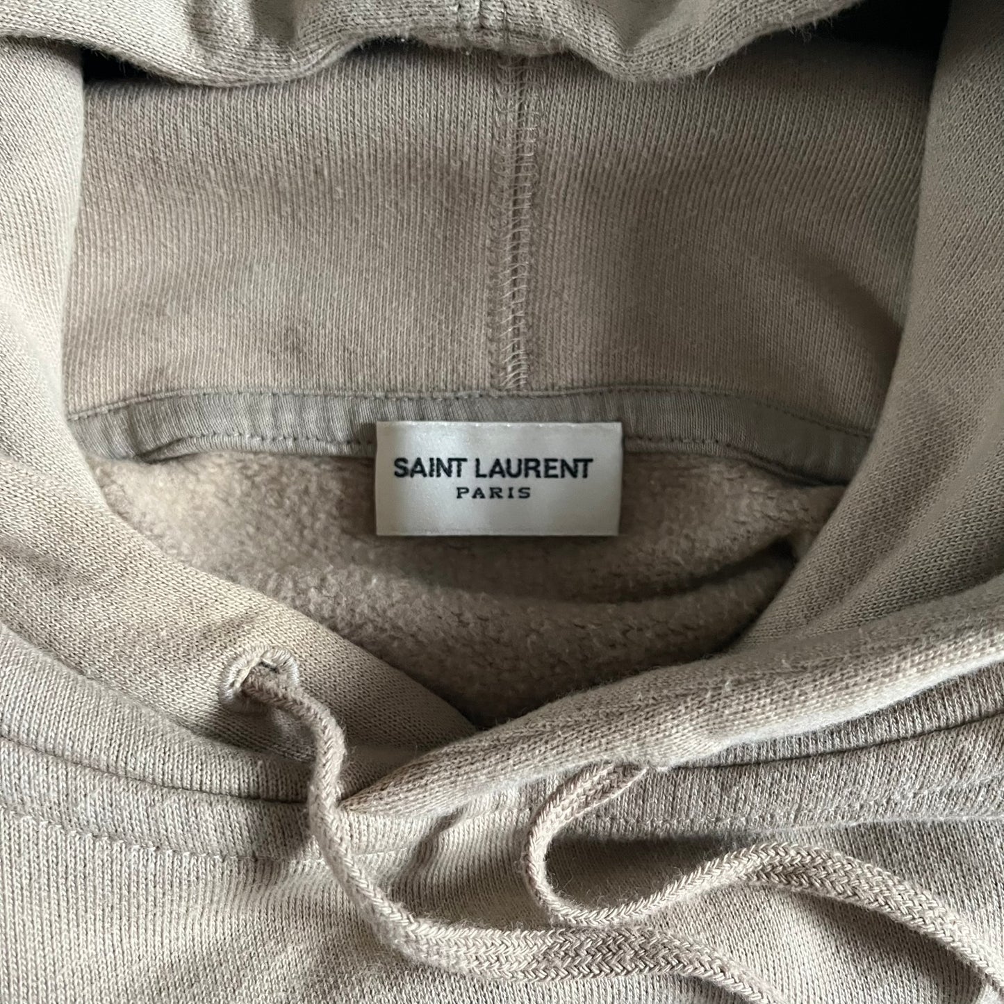 Crafted in France from 100% cotton, this Saint Laurent Beige Hoodie features a minimalist aesthetic with the brand’s signature small Saint Laurent chest logo and red square detail. Designed with a classic front pocket and clean drawstrings without metal tips, it delivers understated Parisian luxury in a neutral tone.