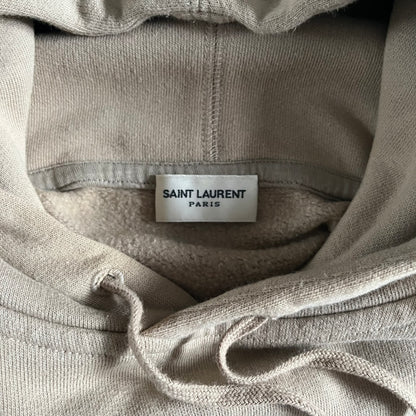 Crafted in France from 100% cotton, this Saint Laurent Beige Hoodie features a minimalist aesthetic with the brand’s signature small Saint Laurent chest logo and red square detail. Designed with a classic front pocket and clean drawstrings without metal tips, it delivers understated Parisian luxury in a neutral tone.