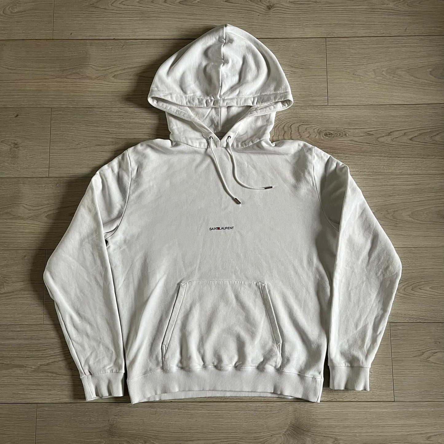 Saint Laurent Small Logo White Hoodie made in France from 100% cotton. Features a minimalist chest logo with “Saint” and “Laurent” separated by a small red square, silver-tipped drawstrings engraved with branding, and a classic front pocket. Designed with the brand’s signature minimalist aesthetic.