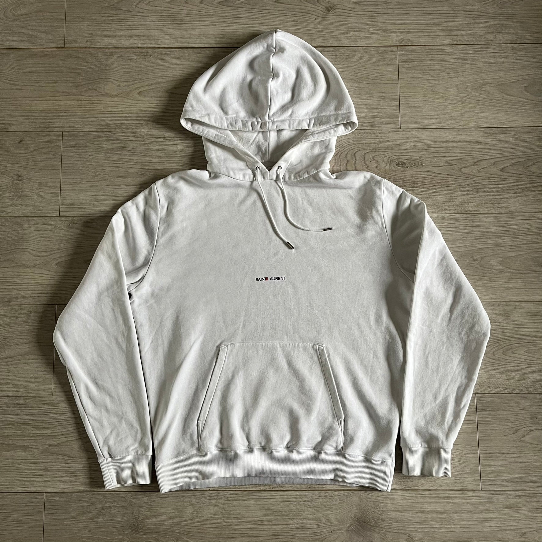 Saint Laurent Small Logo White Hoodie made in France from 100% cotton. Features a minimalist chest logo with “Saint” and “Laurent” separated by a small red square, silver-tipped drawstrings engraved with branding, and a classic front pocket. Designed with the brand’s signature minimalist aesthetic.