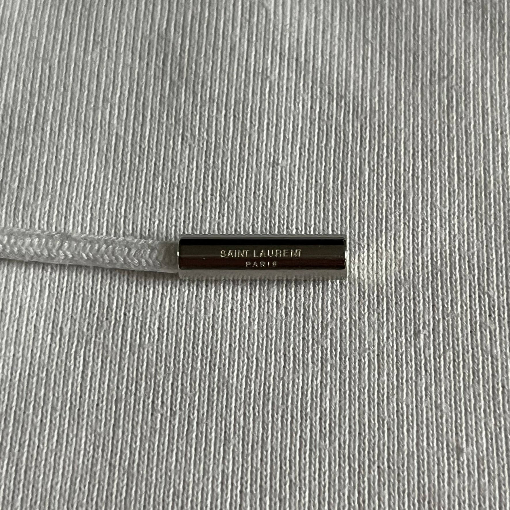 Saint Laurent Small Logo White Hoodie made in France from 100% cotton. Features a minimalist chest logo with “Saint” and “Laurent” separated by a small red square, silver-tipped drawstrings engraved with branding, and a classic front pocket. Designed with the brand’s signature minimalist aesthetic.