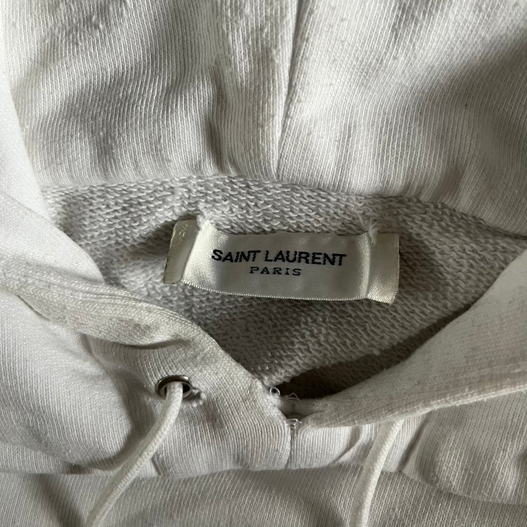 Saint Laurent Small Logo White Hoodie made in France from 100% cotton. Features a minimalist chest logo with “Saint” and “Laurent” separated by a small red square, silver-tipped drawstrings engraved with branding, and a classic front pocket. Designed with the brand’s signature minimalist aesthetic.