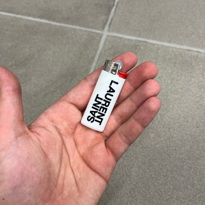 Saint Laurent White Logo Lighter is a white BIC gas lighter featuring black Saint Laurent branding, with “Saint” printed above and “Laurent” mirrored underneath. It is an in-store-only item made in Spain.
