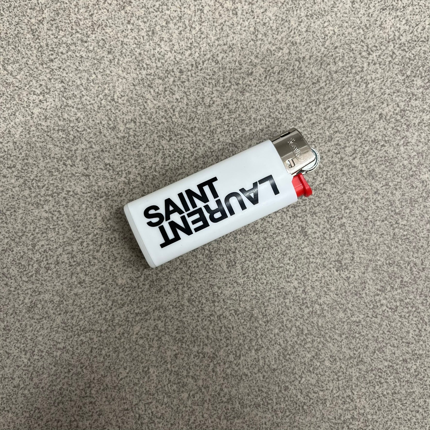 Saint Laurent White Logo Lighter is a white BIC gas lighter featuring black Saint Laurent branding, with “Saint” printed above and “Laurent” mirrored underneath. It is an in-store-only item made in Spain.
