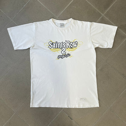 Saints Row 2 x PLNY Textylia 2008 White Promo Tee is a rare Poland-exclusive promotional shirt released for Saints Row 2 in collaboration with Polish brand PLNY Textylia, limited to a numbered run (this piece numbered #00059). The front features a black and white Saints Row 2 logo over yellow tribal graphics with a smaller PLNY Textylia logo below, plus a patch displaying both brand logos, while the back carries a large yellow dot/square-style graphic of the game's cover character.
