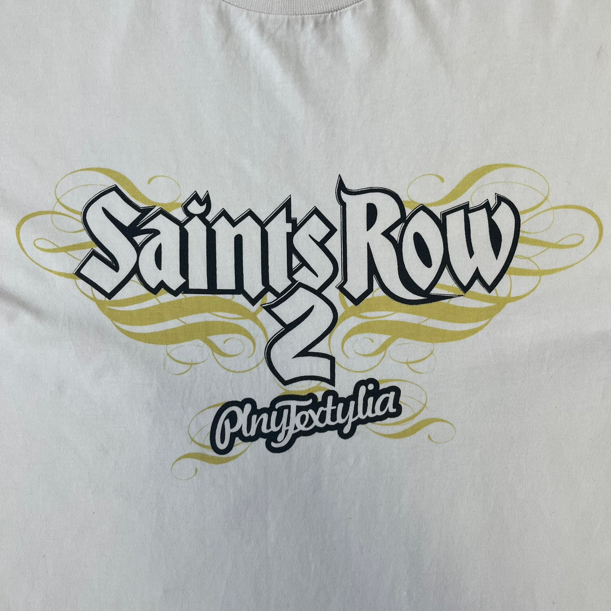 Saints Row 2 x PLNY Textylia 2008 White Promo Tee is a rare Poland-exclusive promotional shirt released for Saints Row 2 in collaboration with Polish brand PLNY Textylia, limited to a numbered run (this piece numbered #00059). The front features a black and white Saints Row 2 logo over yellow tribal graphics with a smaller PLNY Textylia logo below, plus a patch displaying both brand logos, while the back carries a large yellow dot/square-style graphic of the game's cover character.